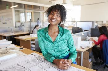 Black female architect leans on desk smiling in busy office