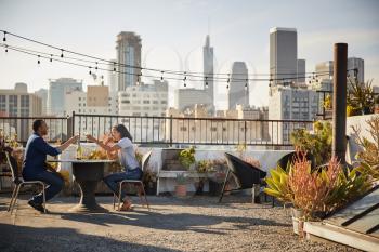 Couple Drinking Wine And Making Toast On Rooftop Terrace With City Skyline In Background