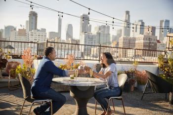 Man Giving Woman Gift As They Celebrate On Rooftop Terrace With City Skyline In Background