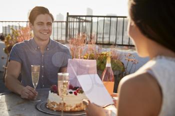 Man Giving Woman Gift And Card As They Celebrate On Rooftop Terrace With City Skyline In Background
