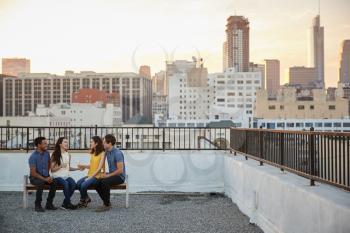 Friends Sitting On Rooftop Terrace Seat With City Skyline In Background