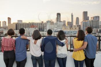 Rear View Of Friends Gathered On Rooftop Terrace Looking Out Over City Skyline