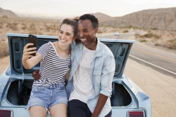 Couple Posing For Selfie Sitting In Trunk Of Classic Car On Road Trip