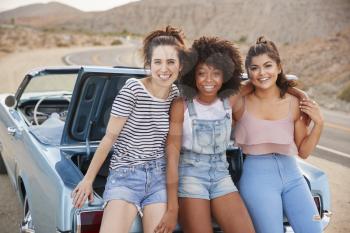 Portrait Of Three Female Friends Sitting In Trunk Of Classic Car On Road Trip