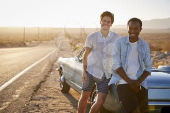 Portrait Of Two Male Friends Enjoying Road Trip Standing Next To Classic Car On Desert Highway
