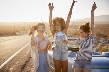 Portrait Of Female Friends Enjoying Road Trip Standing Next To Classic Car On Desert Highway