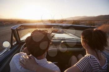 Rear View Of Couple On Road Trip Driving Classic Convertible Car Towards Sunset