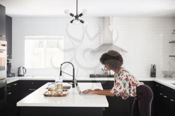 Woman At Home Standing At Kitchen Island Whilst Using Laptop