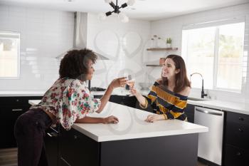 Two Female Friends Making Toast As They Drink Wine At Home Standing By Kitchen Island