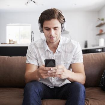 Man Wearing Wireless Headphones Sitting On Sofa At Home Streaming From Mobile Phone