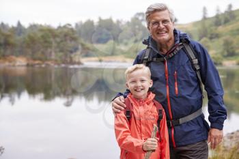 Grandfather and grandson standing together on the shore of a lake smiling to camera, close up, Lake District, UK