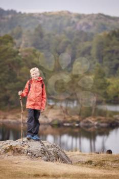A boy standing on a rock by a lake holding a stick, smiling to camera, Lake District, UK, vertical