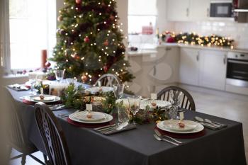 Christmas table setting with bauble name card holders arranged on plates and green and red decorations with Christmas tree and kitchen in the background