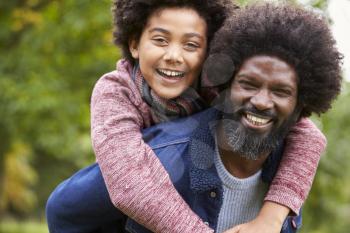 Black man piggybacking his pre-teen son in the park, both smiling to camera, close up