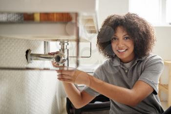 Young black female plumber sitting on the floor fixing a bathroom sink, looking to camera