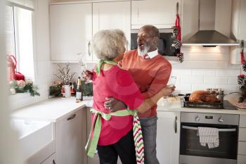 Happy mature black couple holding champagne glasses, laughing and embracing in the kitchen while preparing meal on Christmas morning, close up