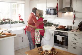 Happy mature black couple holding champagne glasses, laughing and embracing in the kitchen while preparing meal on Christmas morning, side view