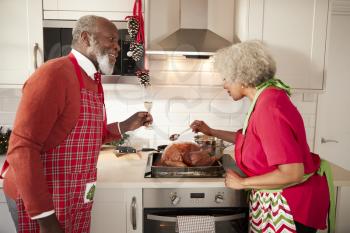 Mature black couple preparing Christmas dinner in their kitchen, man raising a glass as his wife bastes the roast turkey
