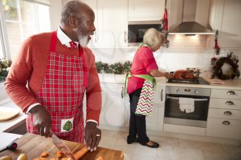 Mature black couple preparing Christmas dinner, man chopping vegetables in the foreground, turning to his partner who is preparing the turkey
