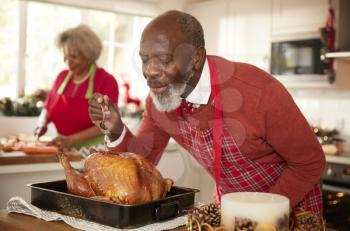 Senior black man basting roast turkey in preparation for Christmas dinner, his wife chopping vegetables in the background, close up, selective focus