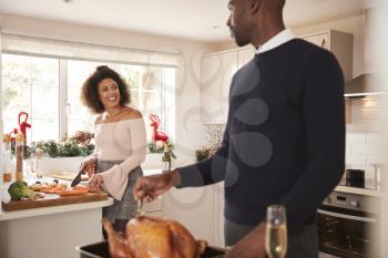 Young adult mixed race couple preparing Christmas dinner together at home, man basting roast turkey in the foreground turning to talk to his partner, front view, close up