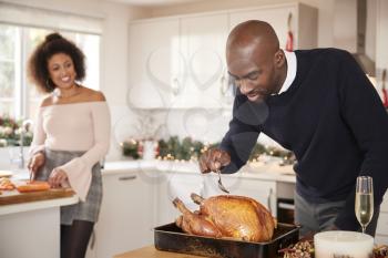 Young adult mixed race couple preparing Christmas dinner together at home, man basting roast turkey in the foreground, front view, close up