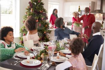 Mixed race, multi generation family gathering in dining room to sit down for Christmas dinner, grandfather holding champagne bottle