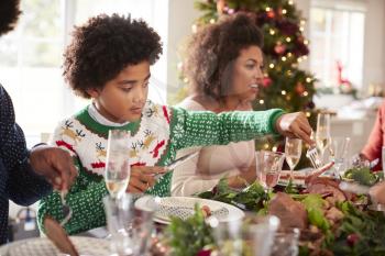 Tween black boy serving himself roast turkey sitting at the Christmas dinner table with his family, side view