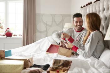 Woman Opening Gift Of Necklace In Bed At Home As Couple Exchange Presents On Christmas Day