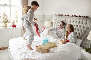 Excited Children Jumping On Parents Bed At Home As Family Open Gifts On Christmas Day