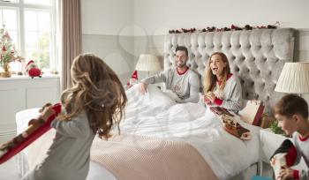 Excited Children Running Into Parents Bedroom At Home With Stockings As Family Open Gifts On Christmas Day