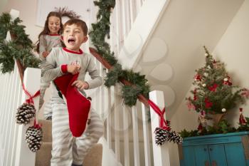 Two Excited Children Wearing Pajamas Running Down Stairs Holding Stockings On Christmas Morning