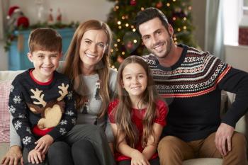 Portrait Of Parents With Children Wearing Festive Jumpers Sitting On Sofa In Lounge At Home On Christmas Day