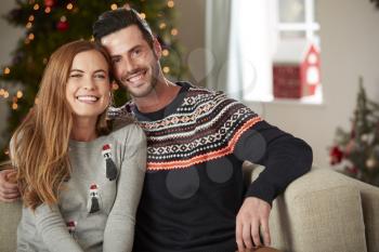 Portrait Of Couple Wearing Festive Jumpers Sitting On Sofa In Lounge At Home On Christmas Day