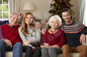 Portrait Of Senior Parents With Adult Offspring Wearing Festive Jumpers Sitting On Sofa In Lounge At Home On Christmas Day