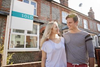 Excited Couple Standing Outside New Home With Sold Sign