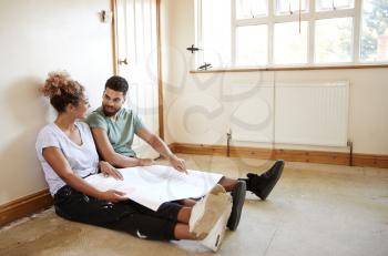 Couple Sitting On Floor Looking At Plans In Empty Room Of New Home