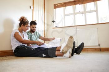 Couple Sitting On Floor Looking At Plans In Empty Room Of New Home