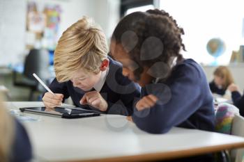 A girl and a boy using a tablet computer and stylus in a primary school class looking closely at the screen, close up