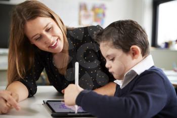 Close up of young female teacher sitting at desk with a Down syndrome schoolboy using a tablet computer in a primary school classroom, smiling, close up, side view