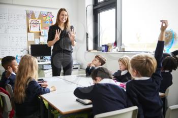 Female primary school teacher standing in a classroom gesturing to  schoolchildren sitting at a table listening, one raising their hand