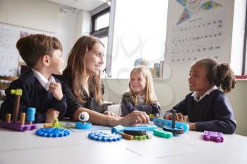 Female teacher and three primary school kids sitting at a table in a classroom working with educational construction toys, close up, low angle