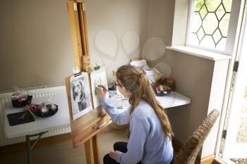 Rear View Of Female Teenage Artist Sitting At Easel Drawing Picture Of Dog From Photograph In Charcoal