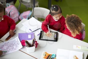 Elevated view of two infant school girls sitting at a table, using a tablet computer and stylus in a classroom