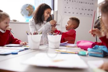 Smiling millennial female infant school teacher sitting at table with kids in a classroom giving pencils to a schoolboy, low angle