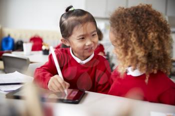 Close up of two kindergarten schoolgirls wearing school uniforms, sitting at a desk in a classroom using a tablet computer and stylus, looking at each other smiling