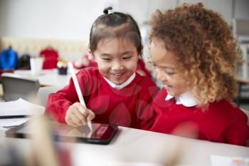 Close up of two kindergarten schoolgirls wearing school uniforms, sitting at a desk in a classroom using a tablet computer and stylus, looking at the screen and smiling