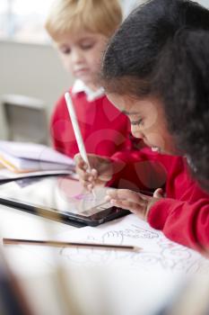 Close up of a schoolgirl sitting at desk next to a boy in an infant school classroom, using a tablet computer and stylus, selective focus, vertical