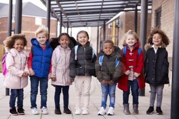 A group of smiling young multi-ethnic school kids wearing coats and carrying schoolbags standing in a row in walkway outside their infant school smiling to camera, full length, front view