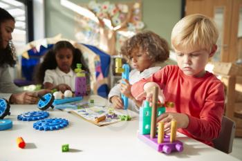 Infant school boy sitting at a table using educational construction toys with his classmates, close up
